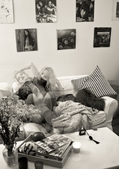 A cozy living room setting with a white sofa covered in a textured blanket and a black-and-white patterned pillow. On the coffee table, there are magazines, a candle, and a small decorative object. A vase with flowers is positioned at the edge of the table. The wall is adorned with several black-and-white posters and photographs, contributing to a vintage ambiance. The double exposure effect gives the image a dreamy, surreal vibe.
