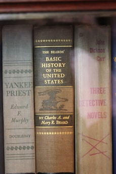 A row of vintage hardcover books with visible spines. Titles include 'Yankee Priest' by Edward F. Murphy, 'Basic History of the United States' by Charles A. and Mary R. Beard, and 'Three Detective Novels' by John Dickson Carr. The books are neatly arranged and display a variety of muted colors and classic typography.