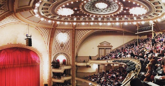 An opulent theater interior with a large audience seated in balconies and rows, adorned with ornate architectural details. The ceiling features intricate designs and chandeliers, while a grand red curtain covers the stage.