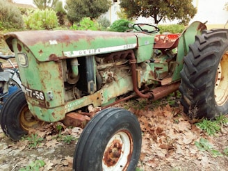 A vintage tractor with rust and wear is parked outdoors surrounded by dry leaves and greenery. The tractor is primarily green with visible rust patches and has large, black tires. The brand name 'John Deere' is faintly visible, and the model appears to be an older version.