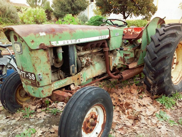 A vintage tractor with rust and wear is parked outdoors surrounded by dry leaves and greenery. The tractor is primarily green with visible rust patches and has large, black tires. The brand name 'John Deere' is faintly visible, and the model appears to be an older version.