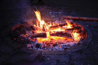 A cozy firepit with burning logs emits bright flames and glowing embers against a dark background. The bricks around the fire form a circular boundary, and some partially burnt wood is scattered on the ground nearby.