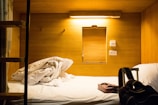 A compact, wooden cabin-style sleeping area featuring a neatly made bed with rumpled sheets. A small window-like opening and two power sockets are visible on the wall. A soft, warm light illuminates the space, creating a cozy atmosphere. On the bed, there's a patterned box and a black backpack with straps.