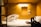 A compact, wooden cabin-style sleeping area featuring a neatly made bed with rumpled sheets. A small window-like opening and two power sockets are visible on the wall. A soft, warm light illuminates the space, creating a cozy atmosphere. On the bed, there's a patterned box and a black backpack with straps.