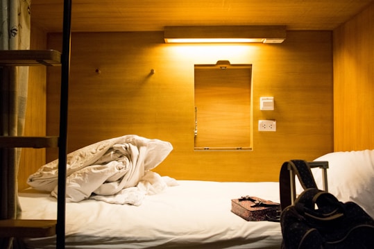 A compact, wooden cabin-style sleeping area featuring a neatly made bed with rumpled sheets. A small window-like opening and two power sockets are visible on the wall. A soft, warm light illuminates the space, creating a cozy atmosphere. On the bed, there's a patterned box and a black backpack with straps.