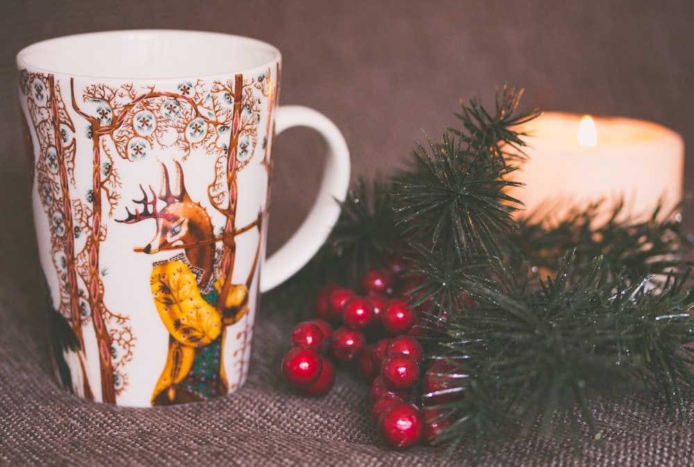 white and brown print ceramic mug near red mistletoe and candle