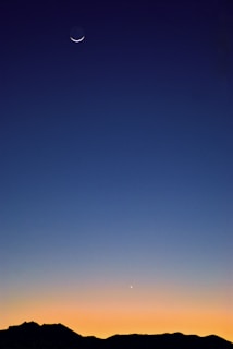 A serene twilight sky with a crescent moon positioned near the top and a bright planet visible below. The gradient shades of blue and orange create a peaceful transition from night to dawn above a silhouette of distant mountains.