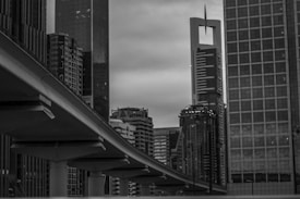 A black and white urban scene featuring a collection of high-rise buildings, including a distinctive skyscraper with a unique cut-out design near the top. In the foreground, there is an elevated roadway or metro track extending across the image, adding a sense of depth and perspective. The skyline is dense with varied architectural styles, highlighting a modern cityscape.