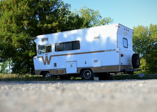 A classic white camper van with a brown stripe is parked on a gravel surface. It has a vintage design, rectangular windows, and a spare tire attached to the back. Trees with lush green leaves are visible in the background, and the sky is clear and blue.