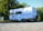 A classic white camper van with a brown stripe is parked on a gravel surface. It has a vintage design, rectangular windows, and a spare tire attached to the back. Trees with lush green leaves are visible in the background, and the sky is clear and blue.