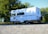 A classic white camper van with a brown stripe is parked on a gravel surface. It has a vintage design, rectangular windows, and a spare tire attached to the back. Trees with lush green leaves are visible in the background, and the sky is clear and blue.