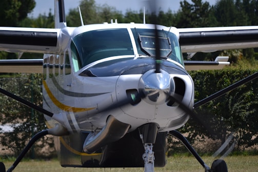 A small propeller airplane is positioned on a grassy runway surrounded by trees. The aircraft's propeller is spinning, creating a blurred effect. The plane has a sleek design with a white, black, and yellow color scheme.