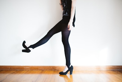 A person is wearing black patterned tights and high-heeled shoes, standing on a wooden floor with one leg slightly raised. The background is a plain, light-colored wall.