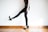 A person is wearing black patterned tights and high-heeled shoes, standing on a wooden floor with one leg slightly raised. The background is a plain, light-colored wall.