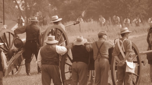 A historical reenactment scene with several individuals dressed in 19th-century attire. They are gathered around a large cannon with wooden wheels, positioned on a grassy field. The people wear wide-brimmed hats, waistcoats, and suspenders. In the background, more figures dressed similarly are visible, adding to the sense of historical ambience.