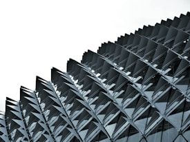 A close-up view of a modern architectural structure, showcasing a pattern of overlapping, triangular metal panels that create a dynamic and geometric facade. The panels appear to be made of a reflective material, likely glass or metal, and are arranged in a repetitive sequence.