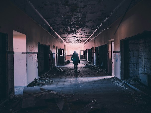 A dimly lit, abandoned corridor with peeling paint and rubble scattered on the floor. A solitary figure is silhouetted against the light at the end of the hallway, creating a sense of mystery and exploration.