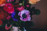 A vibrant bouquet of flowers featuring an array of colors, including deep pinks, purples, blues, and oranges. The flowers appear fresh and are arranged closely together with green leaves providing contrast.