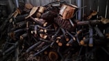 A large stack of cut firewood arranged against a wooden fence. The logs vary in size, with some being larger blocks and others being smaller branches. The wood shows natural textures and has been cut with visible tree rings on the ends.