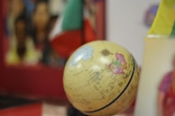A globe displaying geographical details is placed in the foreground. The background is blurred, showing vibrant colors and indistinct objects. Flags are partially visible to the right, adding a multicultural aspect.
