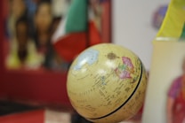 A globe displaying geographical details is placed in the foreground. The background is blurred, showing vibrant colors and indistinct objects. Flags are partially visible to the right, adding a multicultural aspect.