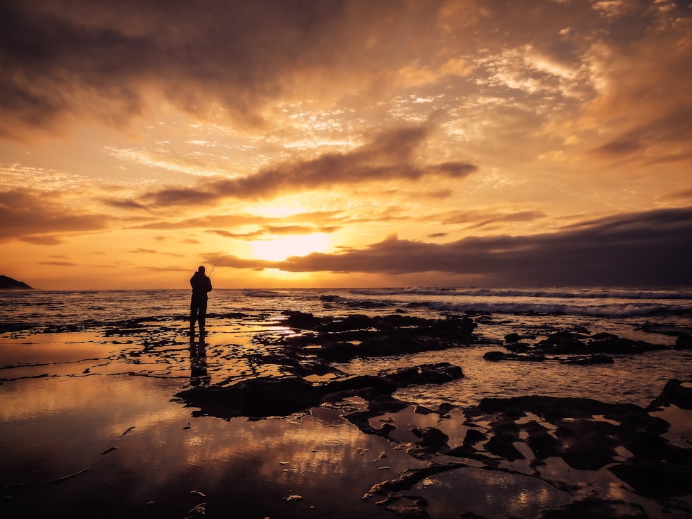 foto silhouette di uomo in piedi sulla spiaggia durante l'ora d'oro