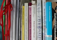 A collection of books tightly packed on a shelf, featuring titles on topics like narrative research, personal stories, and human sciences. Some books are in English, while others are in Finnish. The spines are various widths and colors, and some books have ribbons hanging out.