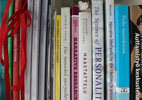 A collection of books tightly packed on a shelf, featuring titles on topics like narrative research, personal stories, and human sciences. Some books are in English, while others are in Finnish. The spines are various widths and colors, and some books have ribbons hanging out.