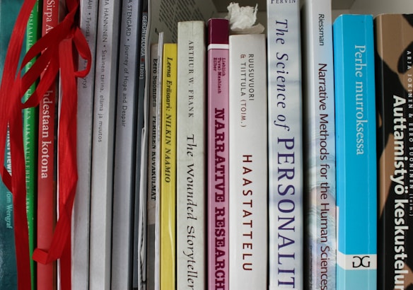 A collection of books tightly packed on a shelf, featuring titles on topics like narrative research, personal stories, and human sciences. Some books are in English, while others are in Finnish. The spines are various widths and colors, and some books have ribbons hanging out.