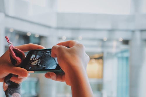A pair of hands holding a smartphone with the camera app open, capturing an image of the surrounding architecture. The background is blurred, highlighting a modern interior space with glass and concrete elements.