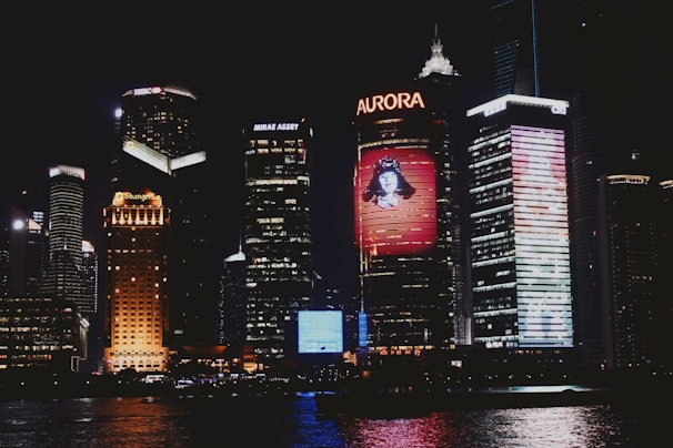 A city skyline illuminated at night with several tall buildings prominently displaying bright advertisements and digital screens. The buildings feature logos such as HSBC, Aurora, and Mirae Asset. Reflections of the lights shimmer on the water in the foreground.