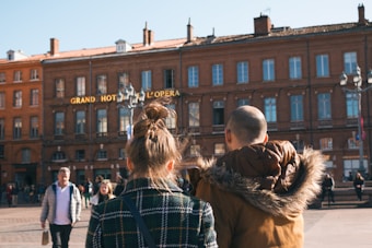 A couple is seen from behind walking through a bustling square with several other pedestrians. The historic facade of a brick building, labeled 'Grand Hotel de l'Opera,' sets a classic European urban backdrop. The scene is lively with people enjoying a sunny day.