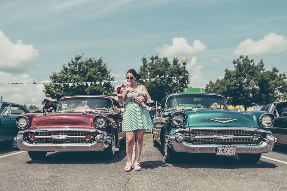 woman carrying child standing between two vintage cars