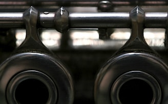 A close-up view of metallic mechanical components with a shiny, reflective surface, featuring two large circular openings and a horizontal rod extending across the top. The image has a slightly blurred background, highlighting the texture and material of the metal.