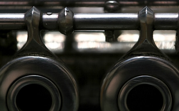 A close-up view of metallic mechanical components with a shiny, reflective surface, featuring two large circular openings and a horizontal rod extending across the top. The image has a slightly blurred background, highlighting the texture and material of the metal.