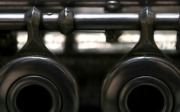 A close-up view of metallic mechanical components with a shiny, reflective surface, featuring two large circular openings and a horizontal rod extending across the top. The image has a slightly blurred background, highlighting the texture and material of the metal.