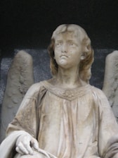 A stone statue depicting an angelic figure with delicate features. The figure has wings behind it and is wearing a flowing robe adorned with subtle floral patterns. The expression on the face is serene and contemplative.