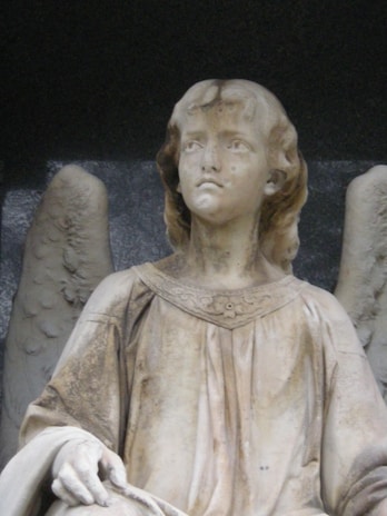 A stone statue depicting an angelic figure with delicate features. The figure has wings behind it and is wearing a flowing robe adorned with subtle floral patterns. The expression on the face is serene and contemplative.