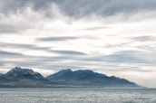 A serene seascape featuring a range of mountains in the background under an overcast sky. The sea appears calm, and the mountains are shrouded in a light mist, enhancing the tranquil atmosphere.