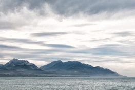 A serene seascape featuring a range of mountains in the background under an overcast sky. The sea appears calm, and the mountains are shrouded in a light mist, enhancing the tranquil atmosphere.