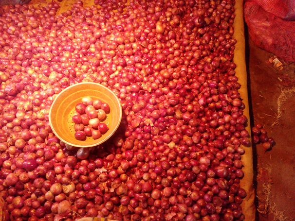 A large number of red onions are spread out on a surface, with an orange bowl containing some onions placed towards the left side. The onions have a shiny appearance due to lighting, and there is a red mesh bag on the upper right corner.