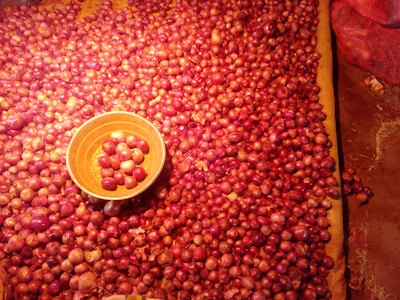 A large number of red onions are spread out on a surface, with an orange bowl containing some onions placed towards the left side. The onions have a shiny appearance due to lighting, and there is a red mesh bag on the upper right corner.