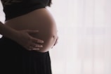 A close-up of a pregnant woman's belly with her hands gently cradling it. She is wearing a black top and a flowing skirt. The background appears soft and light, possibly sheer curtains diffusing sunlight.