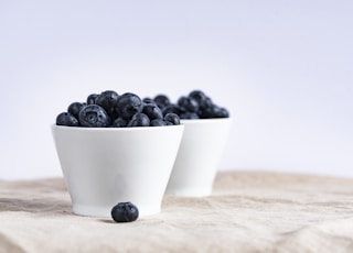 two white ceramic cup filled with blueberries fruit place on brown textile