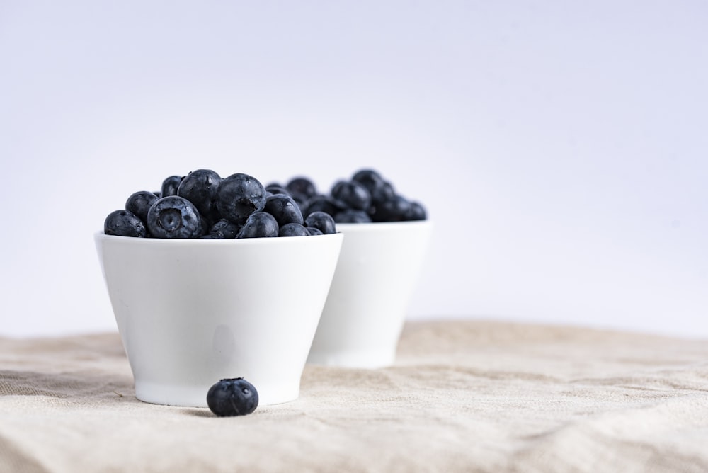 two white ceramic cup filled with blueberries fruit place on brown textile