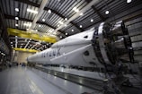 A large rocket is stationed inside a brightly lit industrial hangar, with visible structural supports and various machinery surrounding it. The yellow overhead crane bears the label 'SpaceX'. The rocket's engines and sections of its body are clearly visible.