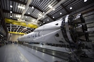 A large rocket is stationed inside a brightly lit industrial hangar, with visible structural supports and various machinery surrounding it. The yellow overhead crane bears the label 'SpaceX'. The rocket's engines and sections of its body are clearly visible.