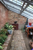 A narrow brick walkway leads through a greenhouse-like space with slanted glass roof panels. On the left, a raised brick planter holds various green plants, while a watering can is nearby. A red cabinet and a black pot are at the far end against a weathered brick wall. On the right, a wooden table is cluttered with various gardening tools, pots, and containers. The atmosphere is rustic and peaceful.