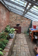 A narrow brick walkway leads through a greenhouse-like space with slanted glass roof panels. On the left, a raised brick planter holds various green plants, while a watering can is nearby. A red cabinet and a black pot are at the far end against a weathered brick wall. On the right, a wooden table is cluttered with various gardening tools, pots, and containers. The atmosphere is rustic and peaceful.