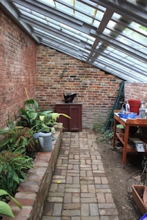 A narrow brick walkway leads through a greenhouse-like space with slanted glass roof panels. On the left, a raised brick planter holds various green plants, while a watering can is nearby. A red cabinet and a black pot are at the far end against a weathered brick wall. On the right, a wooden table is cluttered with various gardening tools, pots, and containers. The atmosphere is rustic and peaceful.